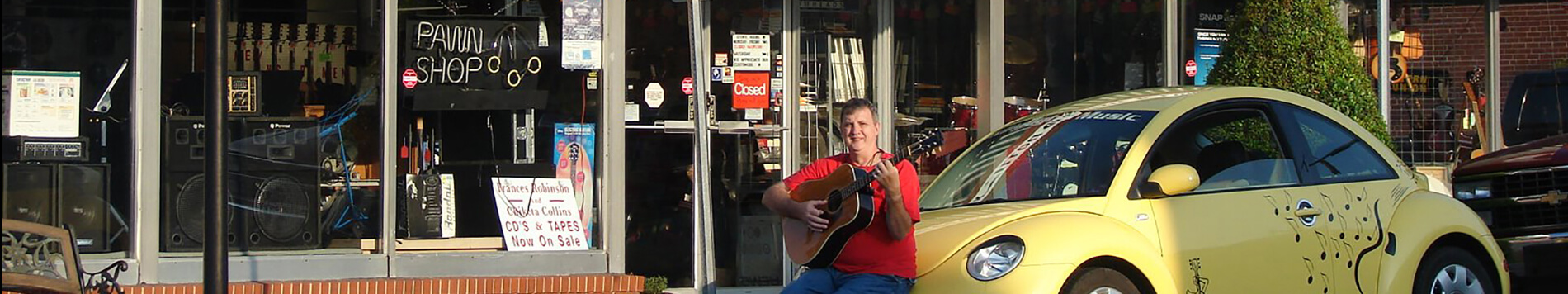 Woody Killough, owner of Killough's Music, playing a guitar outside the store sitting on his yellow vw beetle car